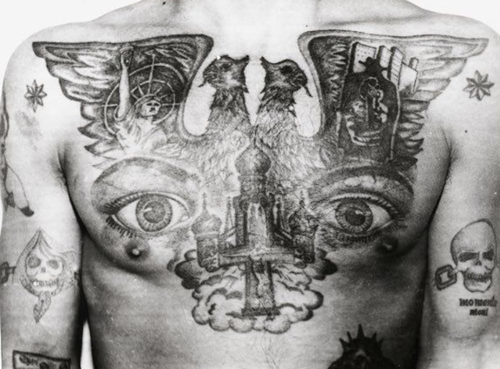 On the arm beneath the skull is the Latin phrase 'Momento mori,' meaning ‘Remember that you will die.' The double-headed eagle is a Russian state symbol that dates back to the 15th century and was used by Peter the Great. In 1993, after the fall of Communism, it replaced the hammer and sickle as the coat of arms of the Russian Federation. This photograph taken in the Soviet period, shows this emblem tattooed as a bold symbol of power and rage against the USSR. It can also be interpreted as ‘Russia for the Russians.' The Statue of Liberty infers a longing for freedom, while the dark character holding a gun denotes a readiness to commit violence and murder. The eyes on the chest signify ‘I can see everything’ and ‘I am watching,' the powerful tattoo of a criminal ‘overseer.' The eight-pointed stars tattooed on the shoulders mark the bearer as an ‘authoritative’ thief.