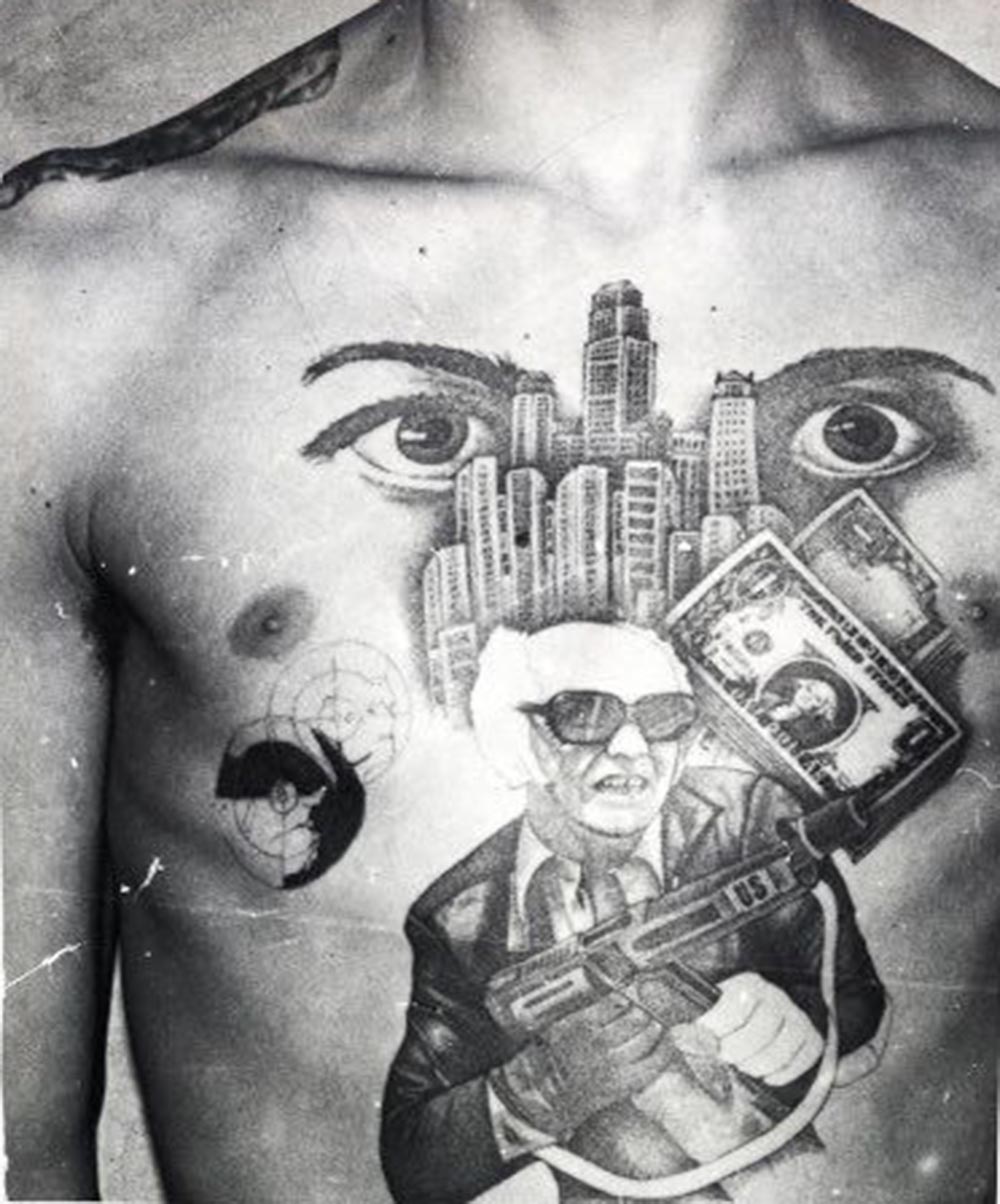 The dollar bills, skyscrapers and machine gun with the initials ‘US’ stamped on it convey this inmate’s love for the American mafia-like lifestyle. The eyes signify ‘I’m watching over you’ (meaning the other inmates in the prison or camp).