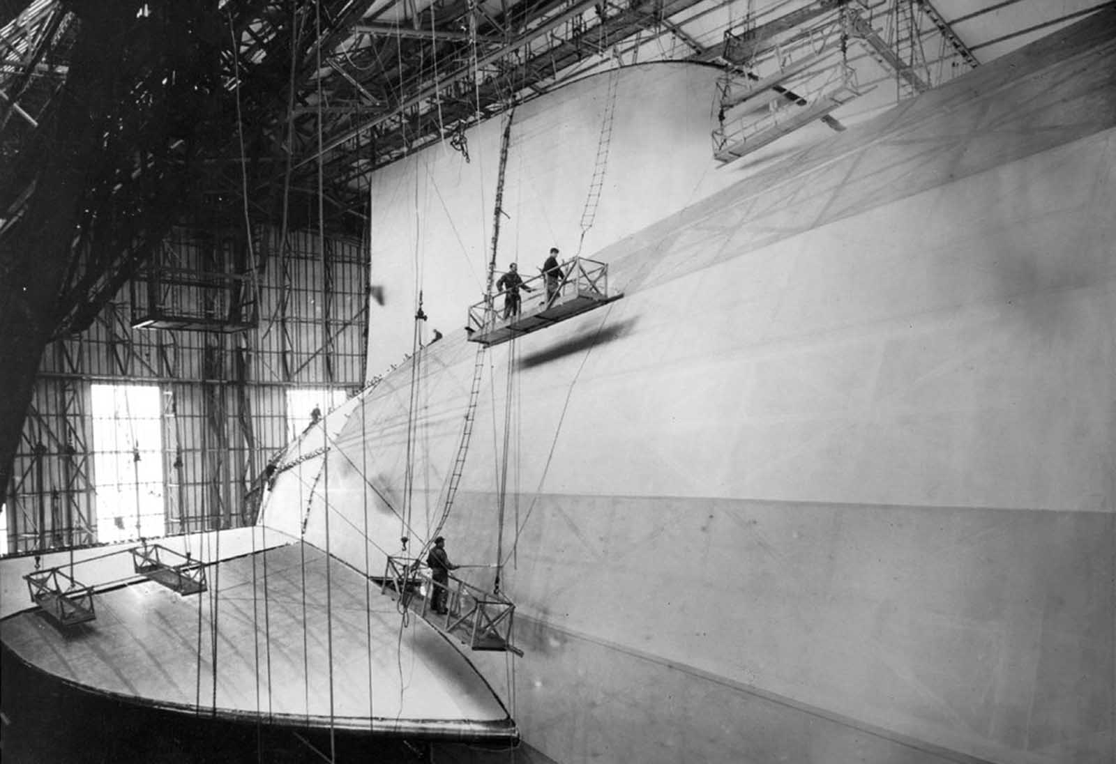 Finishing touches are applied to the A/S Hindenburg in the huge German construction hangar at Friedrichshafen. Workmen, dwarfed in comparison with the ship's huge tail surfaces, are chemically treating the fabric covering the huge hull.