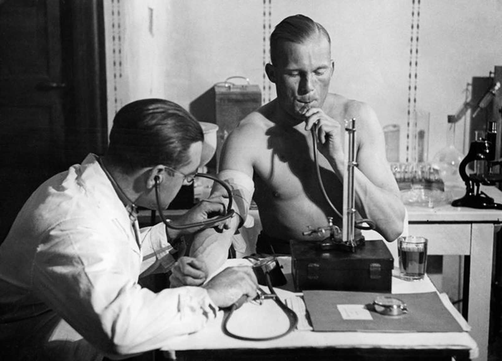 Lieutenant Radtke presses air into his lungs in a constant height with a mercury column, while the doctor checks his blood pressure, circa 1932.
