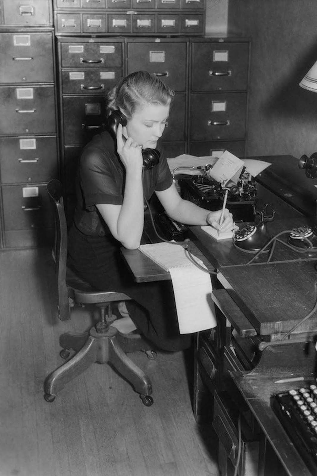 An American secretary sits at a desk behind a typewriter, on the phone taking messages. She wears black, and her blouse is semi-sheer. Her hair is worn neck-length with a permanent wave. 1930.