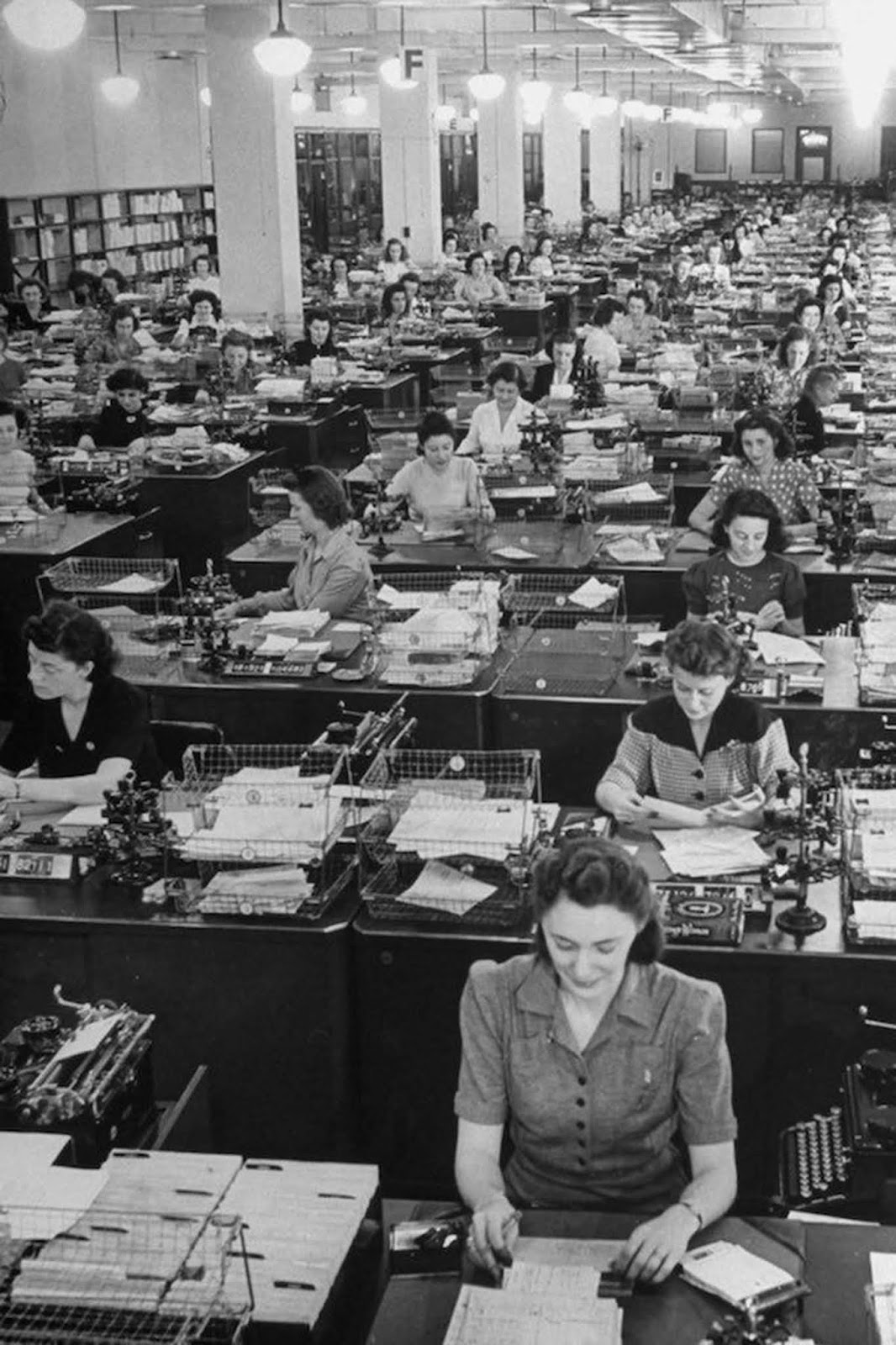 325 American women working to fill mail orders in the office of the Book of the Month Club. The women wear a wide variety of blouses and dresses, including decorative patterns and polka dots, though most have V-necks. Some wear brooches. They typically wear shoulder-length hair; some, as the woman at the front, use hair nets. 1942.