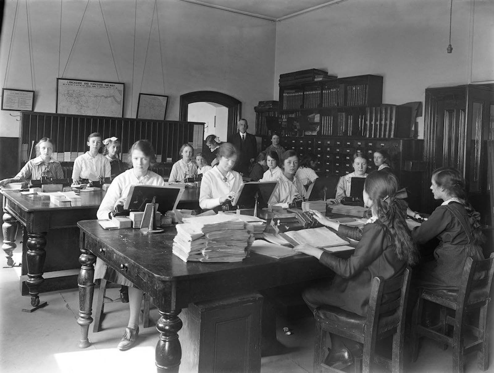 Office at the railway works in Horwich, Lancashire, England. During the First World War many male rail workers joined the army, so women were employed in their place, in a variety of roles including blacksmiths, welders and electricians. Most of the women in this office wear white blouses, but the blouses are more closely fitting than in the previous picture. Their hair is still worn up, though now more loosely, and one woman — at the card cabinets — has shoulder-length hair worn down, while another wears long braids. At least two younger women also have white bows in their hair. The woman in the left foreground shows a shorter skirt length and a leather shoe with a golfing-style flap. 1917.