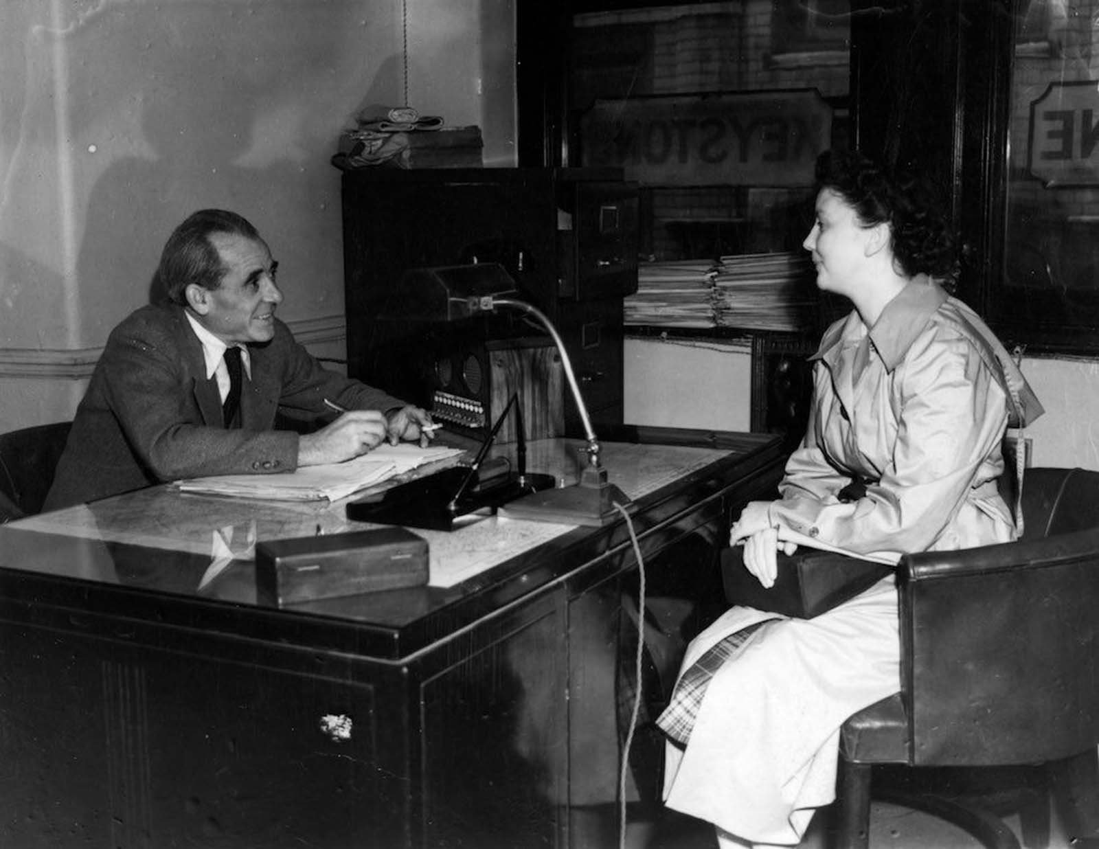 A young woman being interviewed for a job in the Keystone photographic agency offices. Her male interviewer wears a tie and large collar, while holding a cigarette. She still wears her raincoat indoors, under which we can spot her plaid skirt. She has long hair, tied up. 1950.