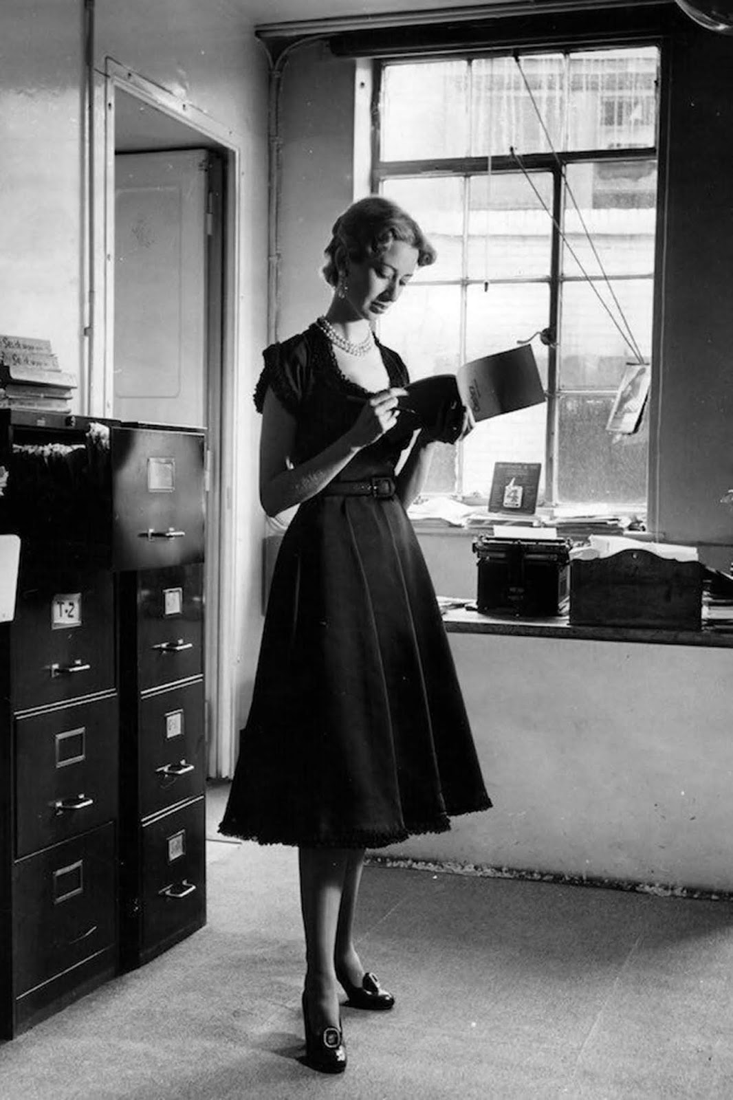 Published in 'Picture Post' with the original caption: 'A woman wearing a black satin dress by Brenner with baby lace around the sleeves, neck and hemline which is not really suitable for the office.' 1950.