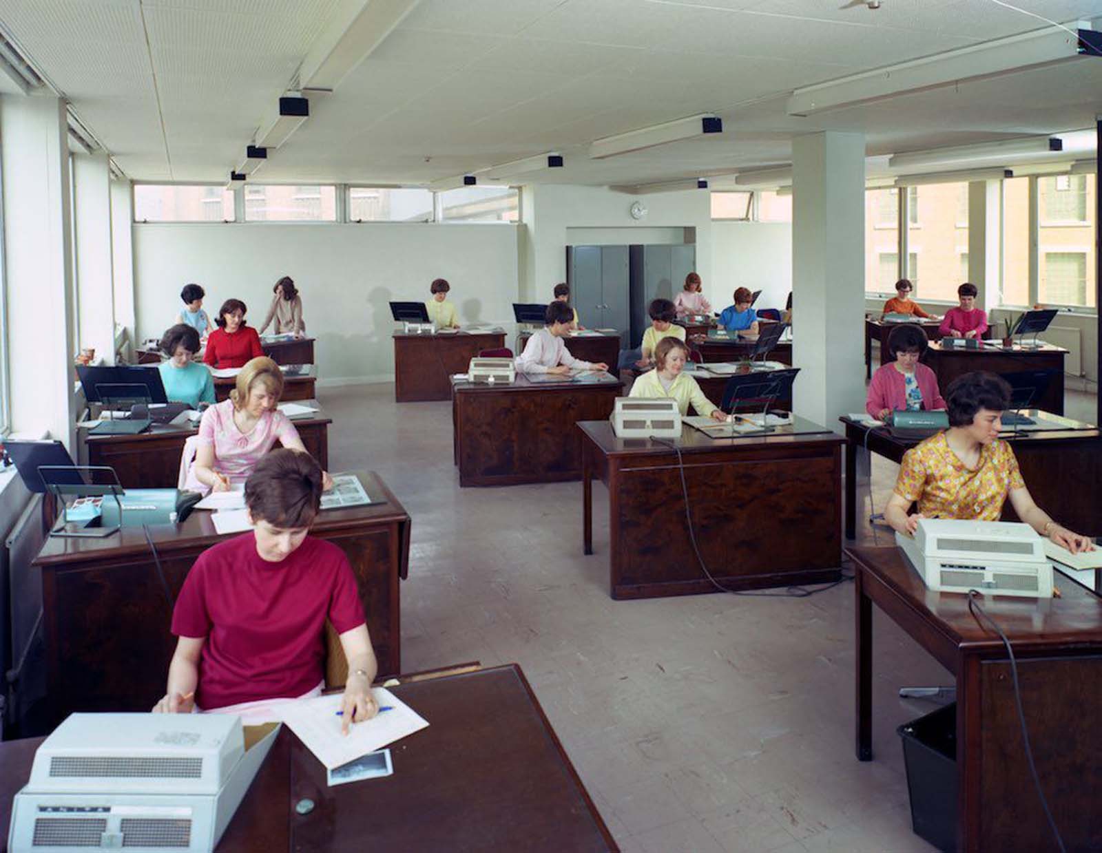 Administration office at Huntsman House, headquarters of Tetley's brewers in Leeds, West Yorkshire. Women workers use Sumlock machines, forerunners of the calculator. They wear clothes in a palette of bright colours. Of the 17 women in the picture, four wear pastel yellow, three wear pastel pink, two wear pastel blue. The women have a wide variety of hair styles, most strongly volumized with hairspray. 1968.