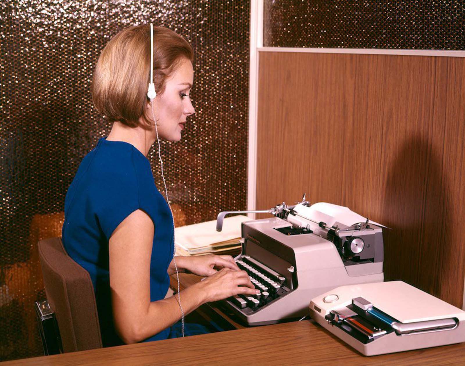 An American secretary typing whilst listening to dictation on her headset. She wears a sleeveless blue top and a blue checked skirt. Her hair is sharply cut and worn chin-length in a brushed bob. 1960s.