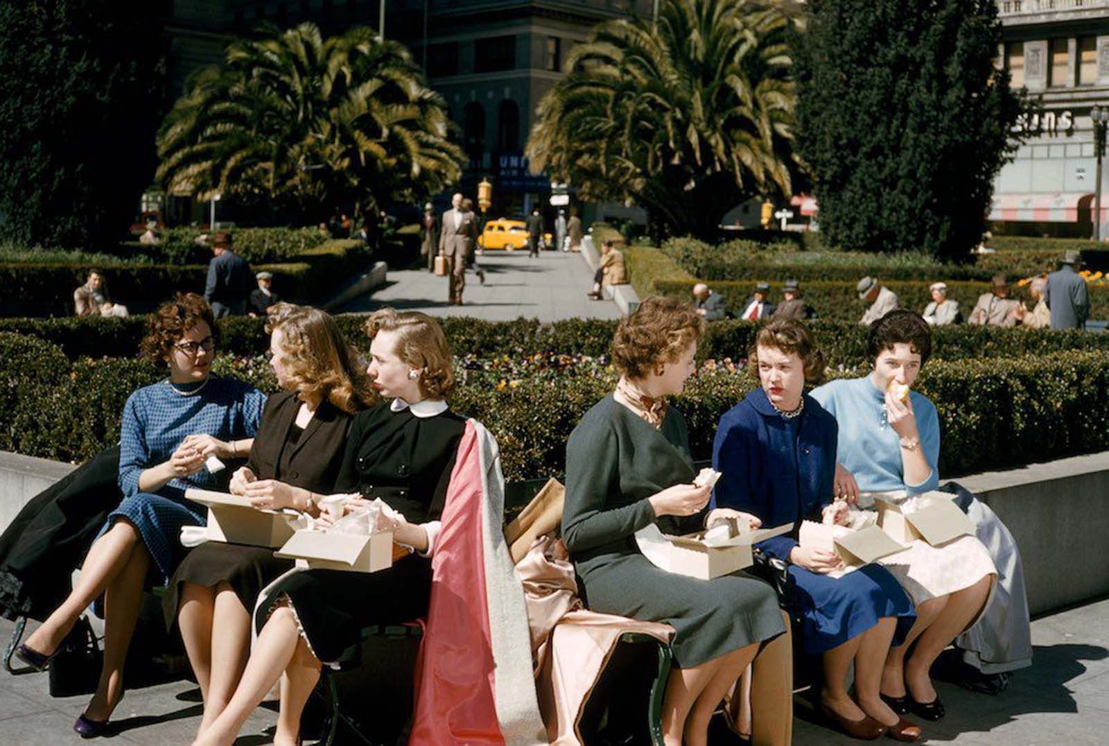 Women office workers eat boxed lunches in Union Square, San Francisco, California. Their suits are formal, in conservative colours. Their hair is worn short, typically parted at the side. All the women appear to be wearing tan stockings or hose. 1956.