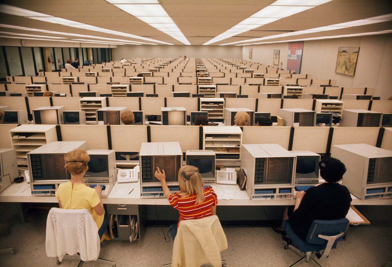 Women operators man computers at Eastern Airlines' reservations center in Miami, Florida. The woman in the centre-front wears a brightly striped top and very naturally styled hair, held loosely in a clip. The woman to the left wears a yellow cotton top and shoes. Cardigans are loosely flung over the backs of their chairs. 1970.