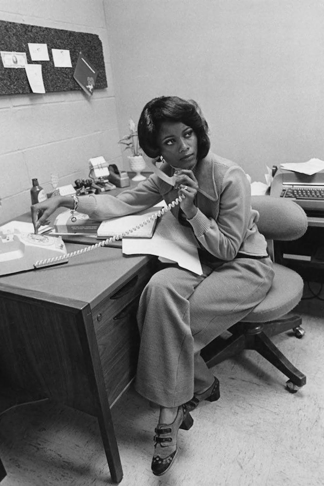 An African-American office worker takes a phone call. She wears a loose-fitting cardigan with a wide collar and wide, loose pants. Her shoes are unusually patterned. She wears a bracelet and ring, and her hair is lightly styled. 1975.