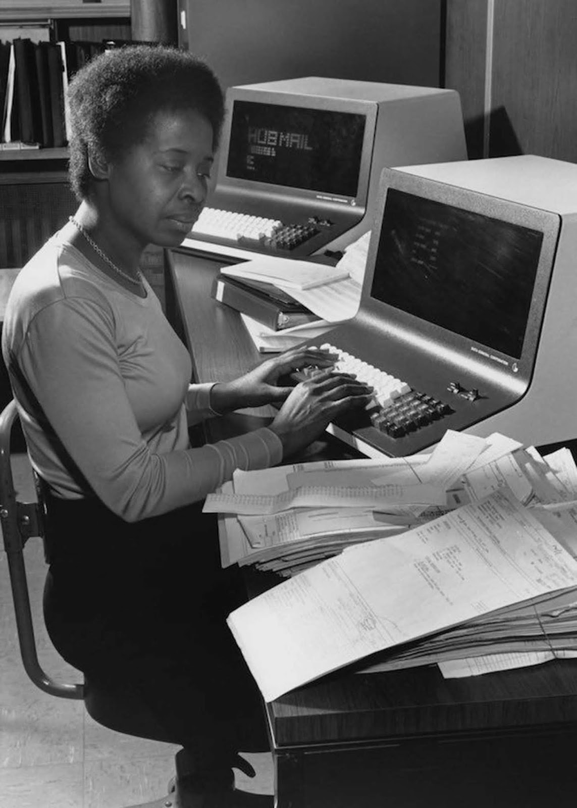 An office worker logs addresses into a Data General computer at a direct-mail company in Boston, Massachusetts. She wears a knit, fitted top; her hair is naturally styled and worn short. 1980.