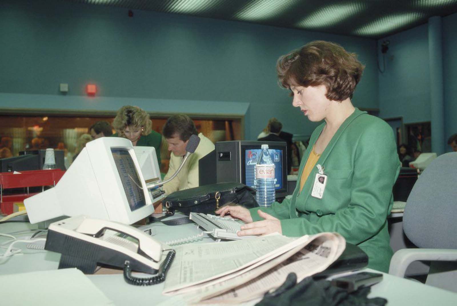 An office worker during the launch of Sky TV in London. She wears a green double-breasted jacket, possibly part of a uniform. Her hair is feathered in layers. In the background another woman wears a similar jacket. Her hair is bleached and waved over large earrings. 1989.