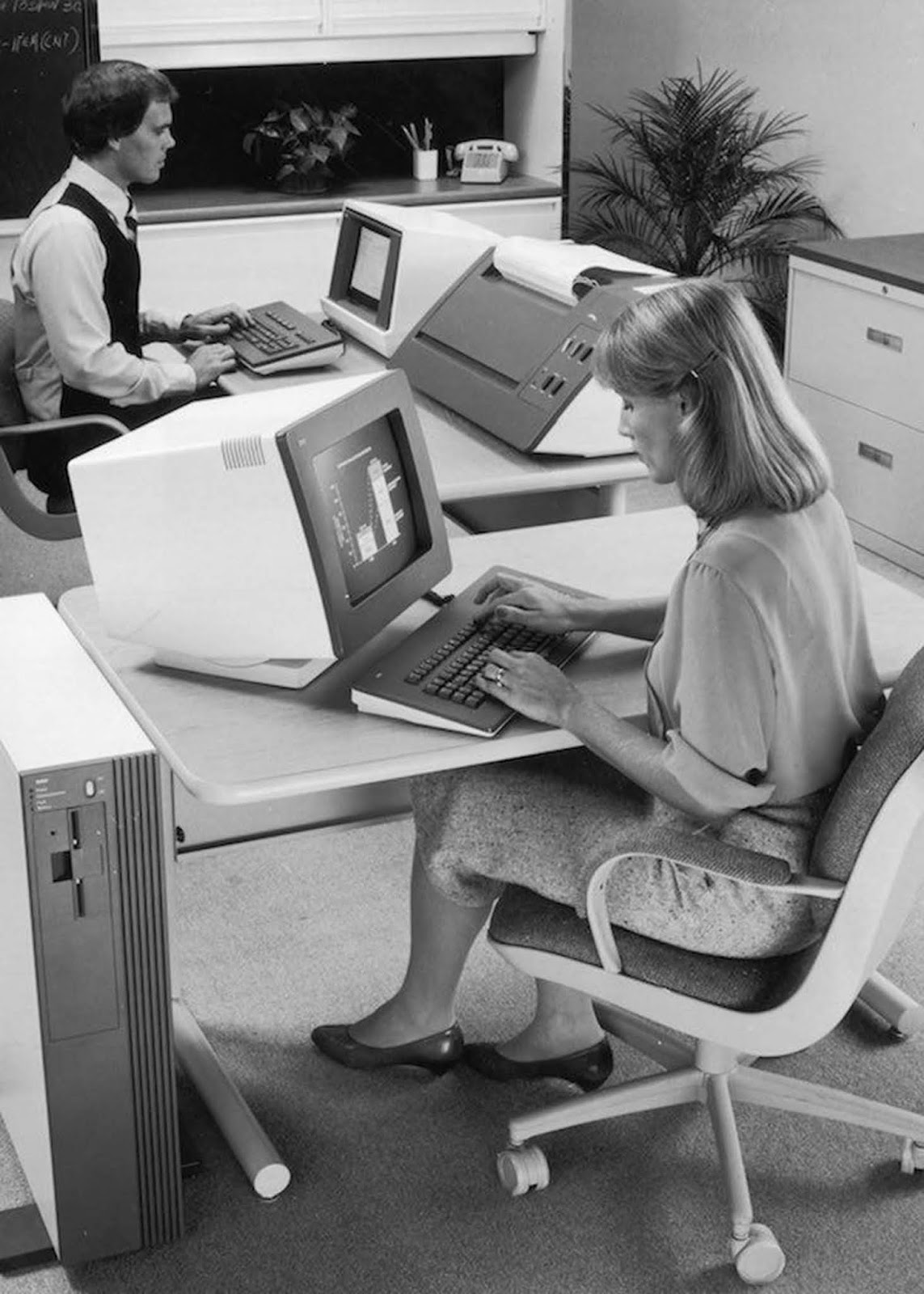 A woman uses a personal computer at the NCR research and development facility in West Columbia, South Carolina. She wears a blouse with rolled sleeves and a formal knee- or calf-length tweed skirt, with court shoes. Her hair is shoulder-length, held away from her face by a slide. 1982.