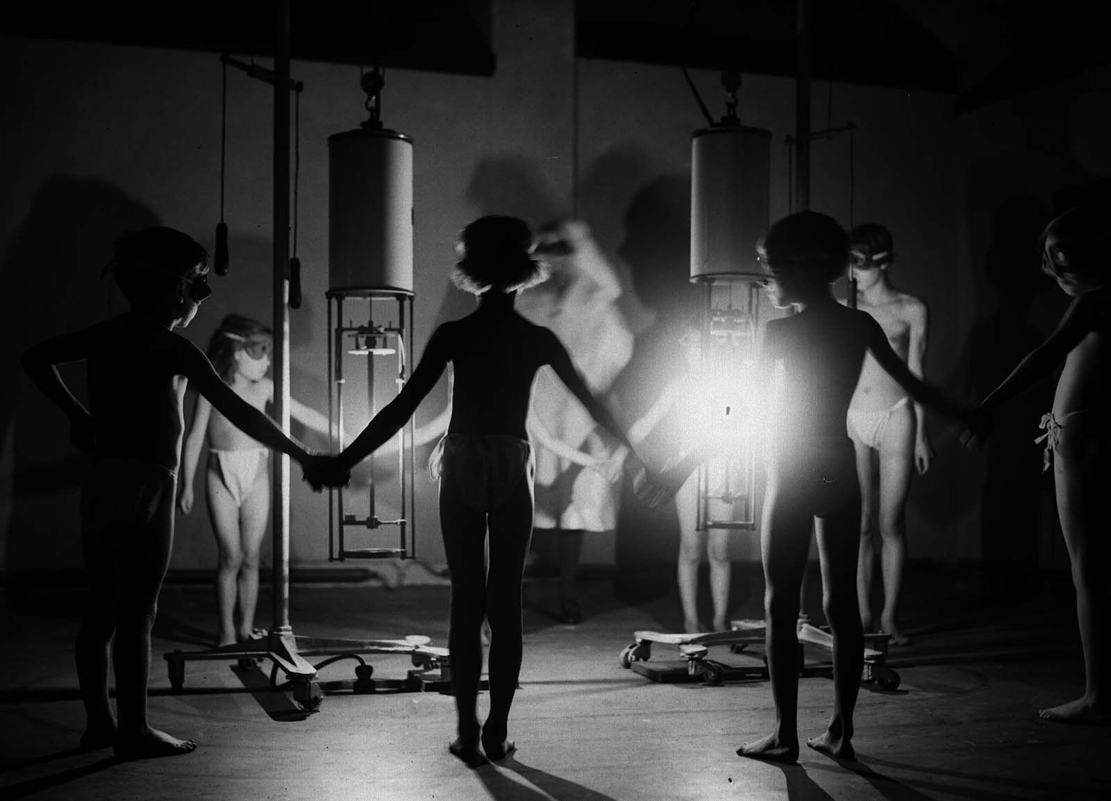 A group of children hold hands around a radiating glow of ultraviolet light at the Institute of Ray Therapy.