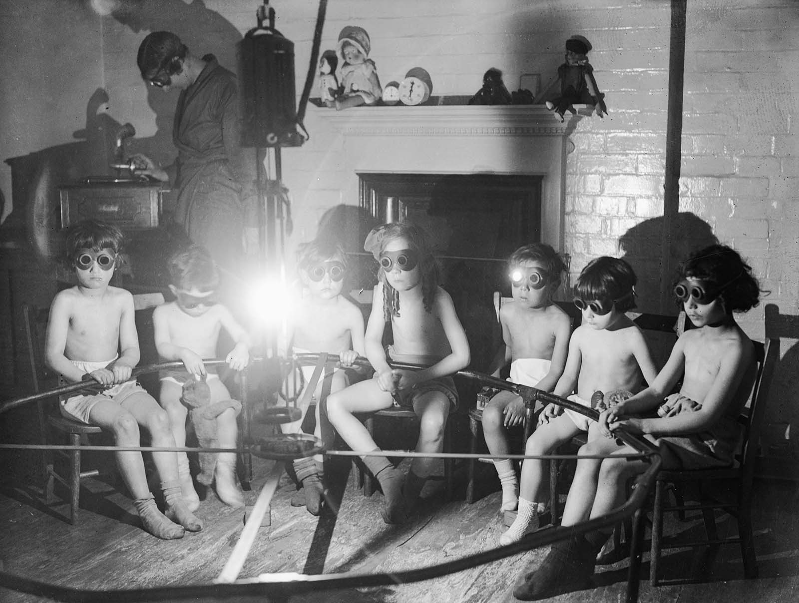 Stripped to the waist and wearing goggles, children sit round a lamp having sun ray treatment while they listen to the gramophone at the East End Mission in Commercial Rd, London. 1931.