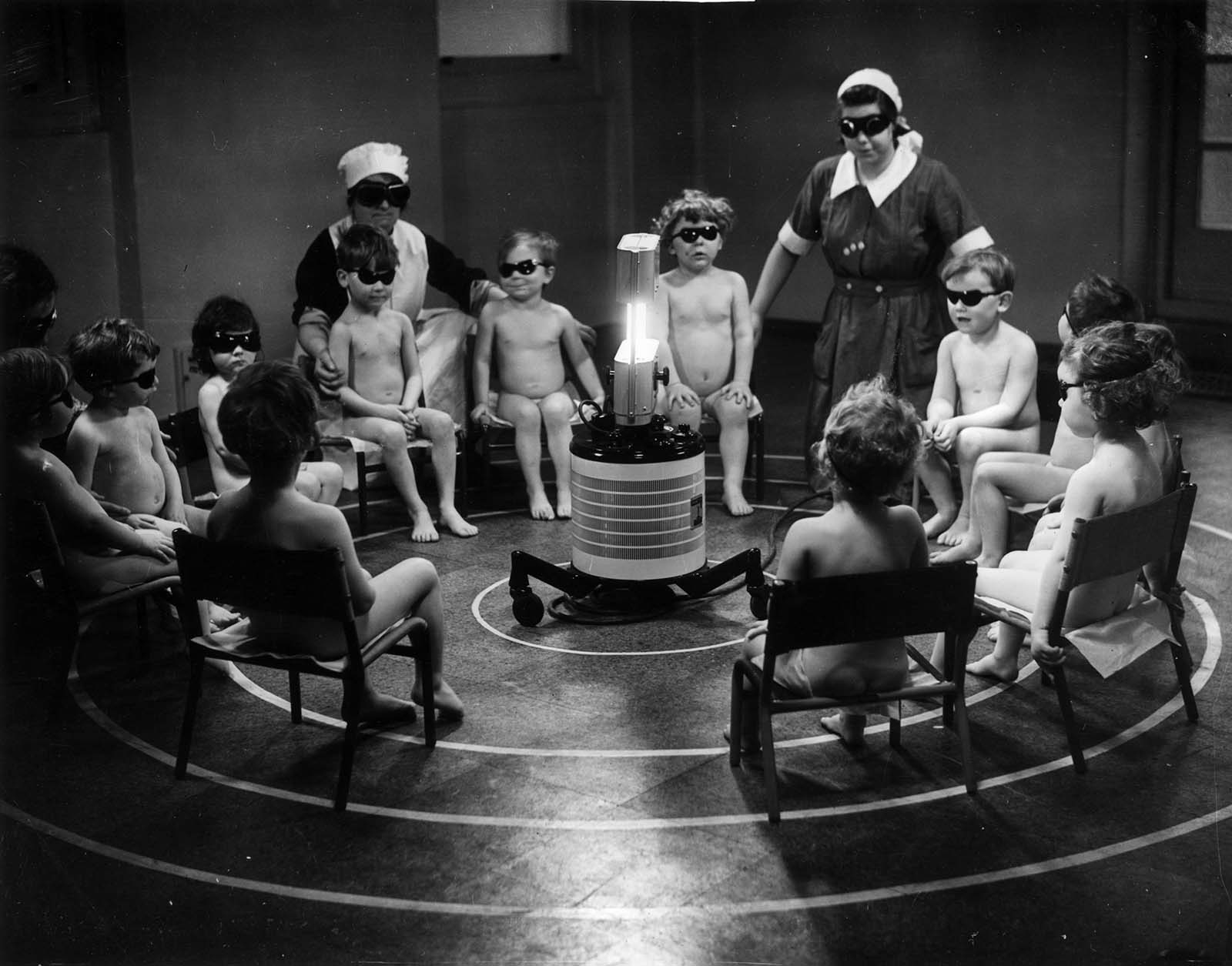 Three nurses and a group of young children sitting around a Centrosol sunray lamp at the Miriam Price Coleman day nursery. 1938.