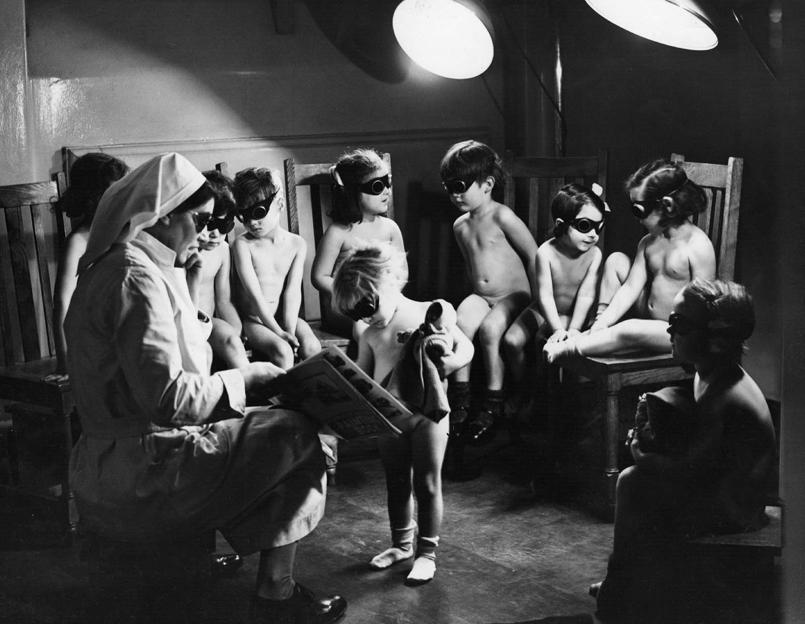 A nurse watching over children in Bermondsey, London, sitting under sun-ray lamps to help make up the deficiency in sunlight and the lack of certain items of food, such as fruit, during the winter months of the Second World War. 1942.