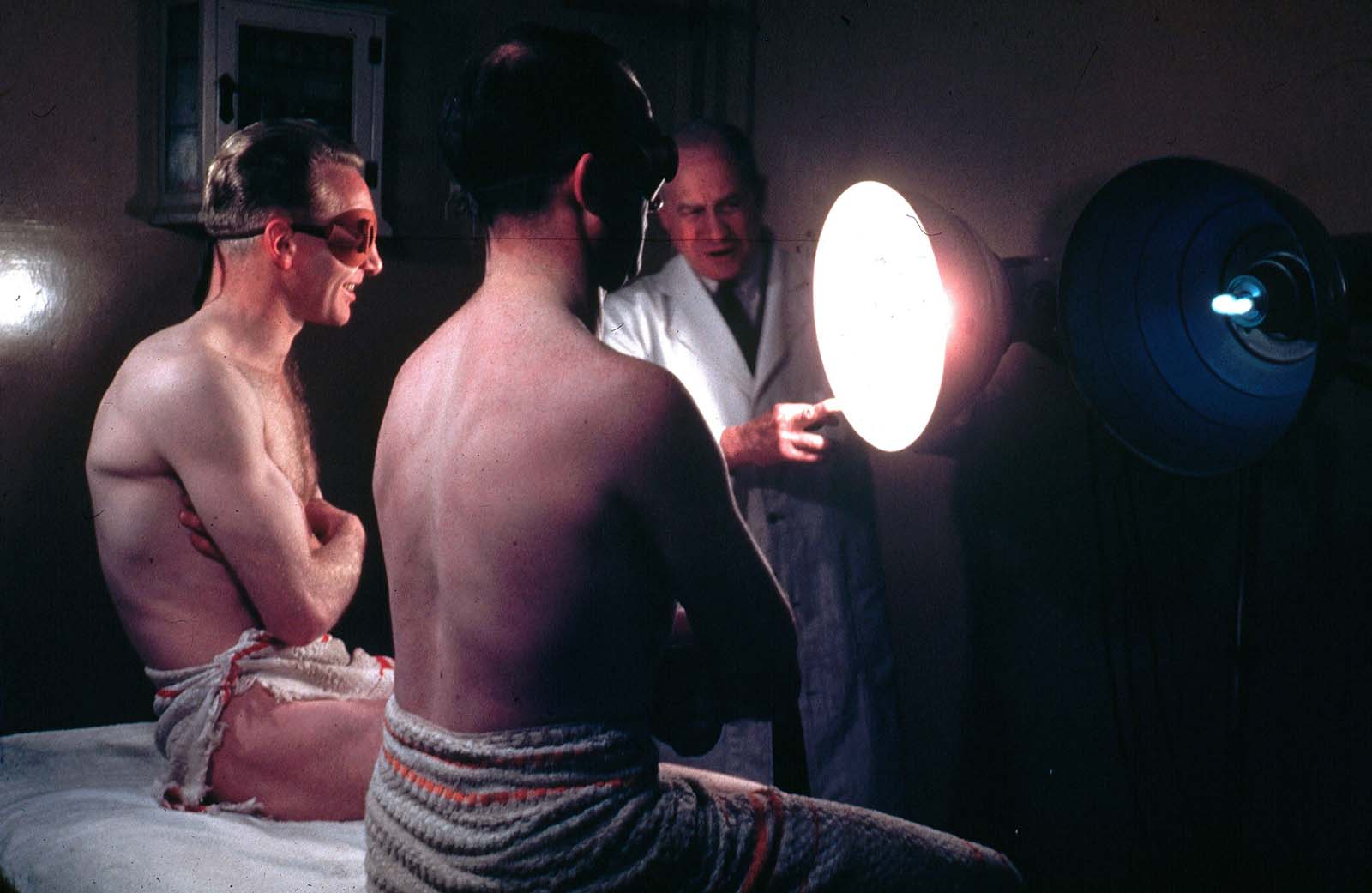 Two men wearing protective eye goggles and towels sitting under sunlamps set up by a doctor. 1950.