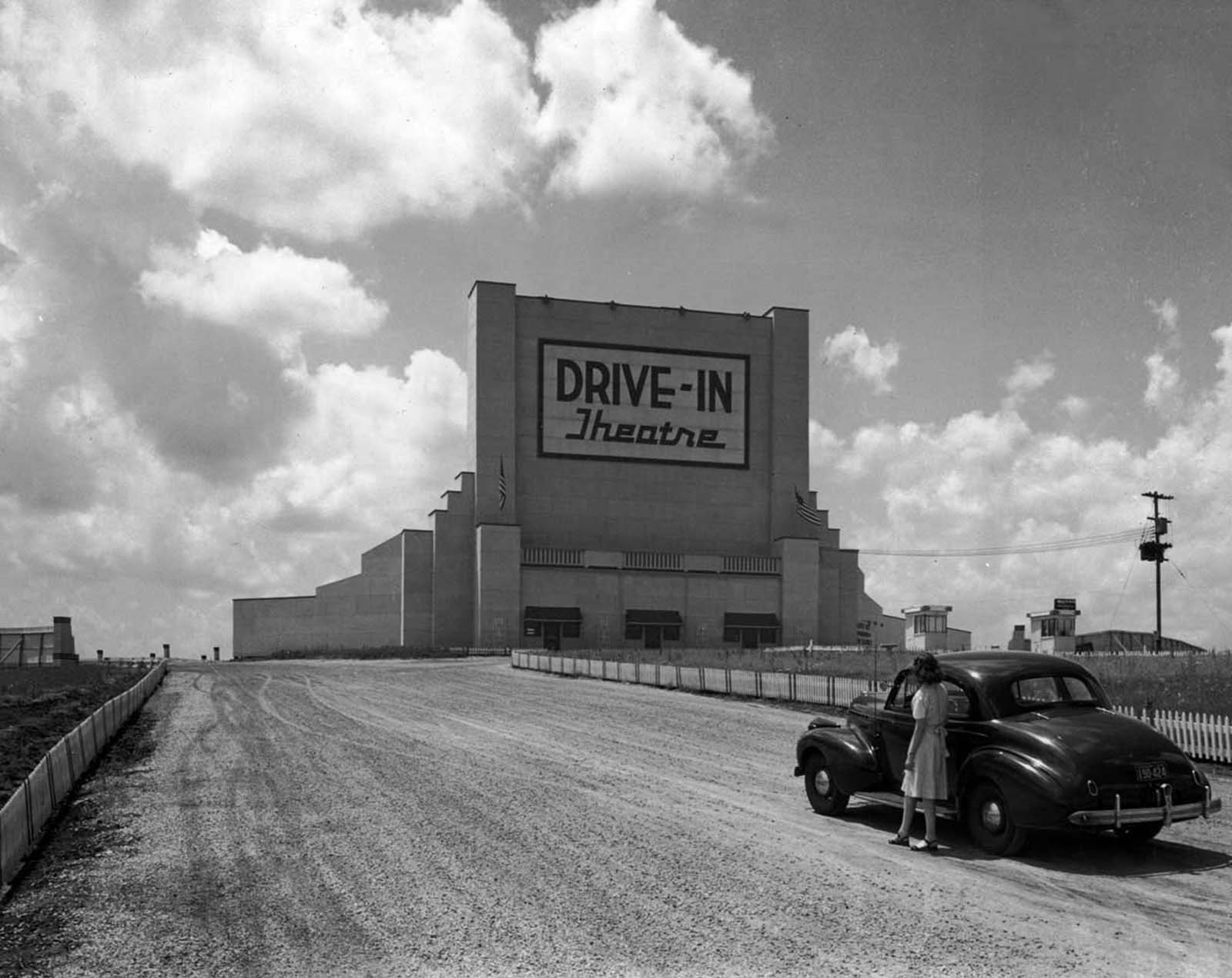 Full-length image of a woman standing next to a parked car in the middle of the road, looking up at a sign that reads, ‘Drive-In Theatre’.