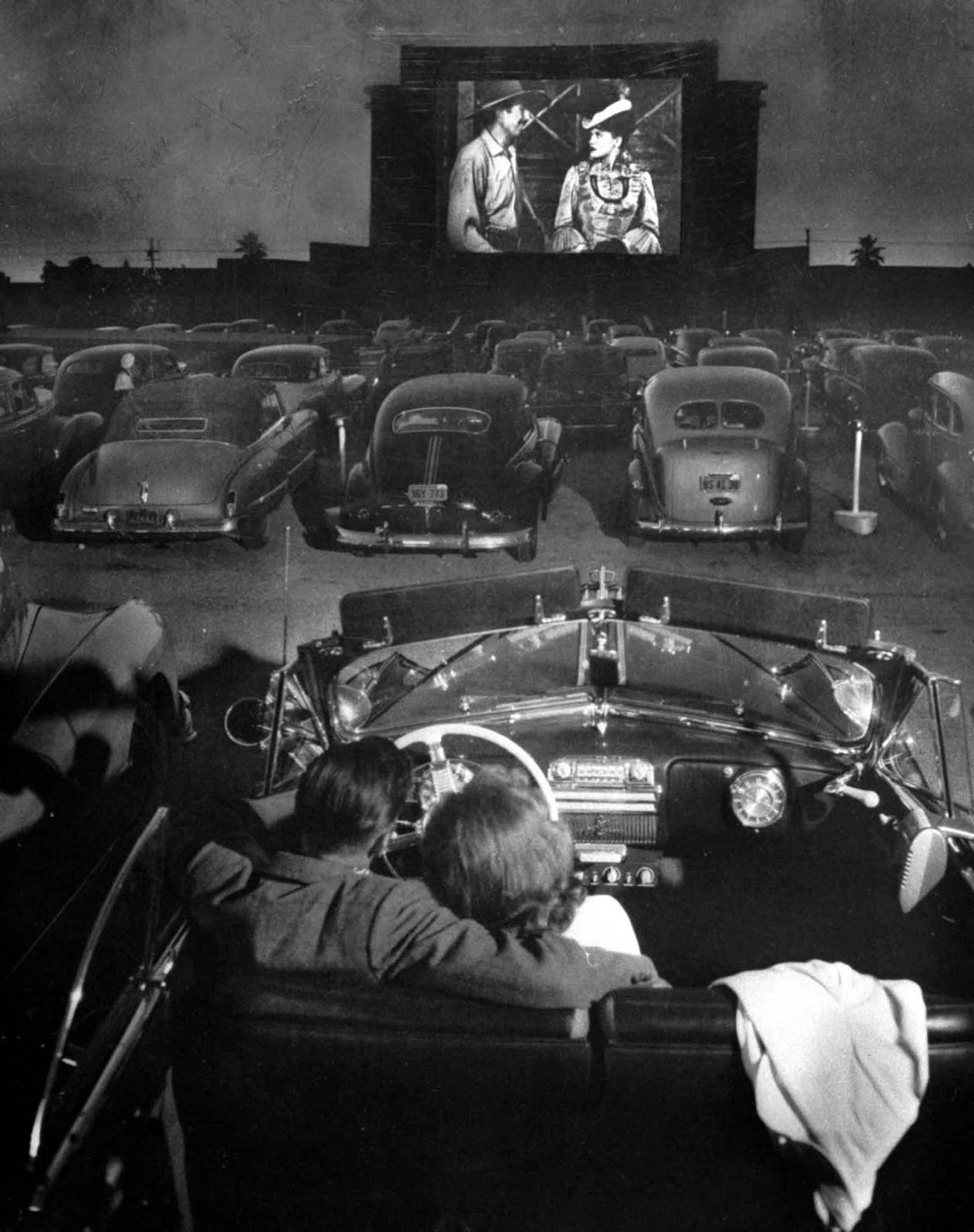 Rear view of young couple snuggling behind the wheel of his convertible as they watch large screen action behind rows of cars at a drive-in movie theater.