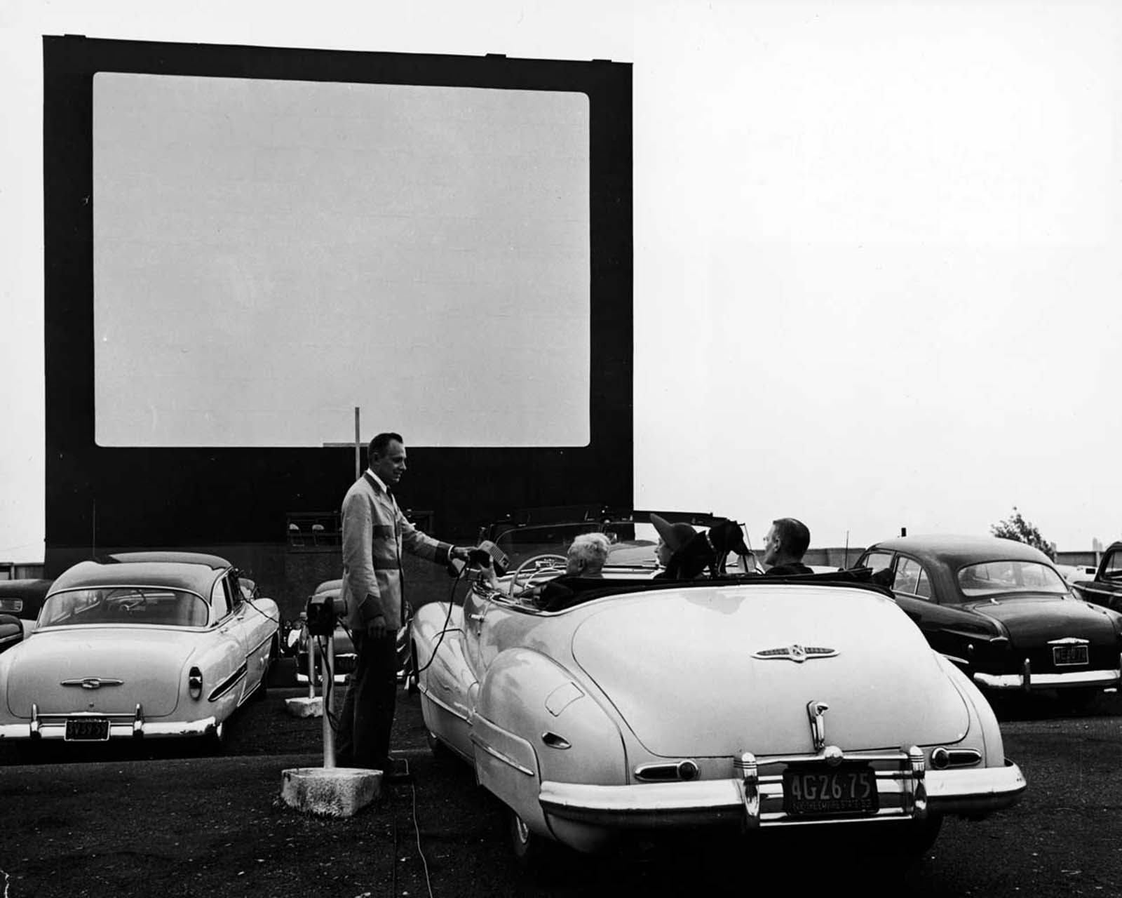 A uniformed drive-in theater attendant hands a clip-on speaker to the driver of a convertible while the car’s other passengers watch, New York, early 1950s.