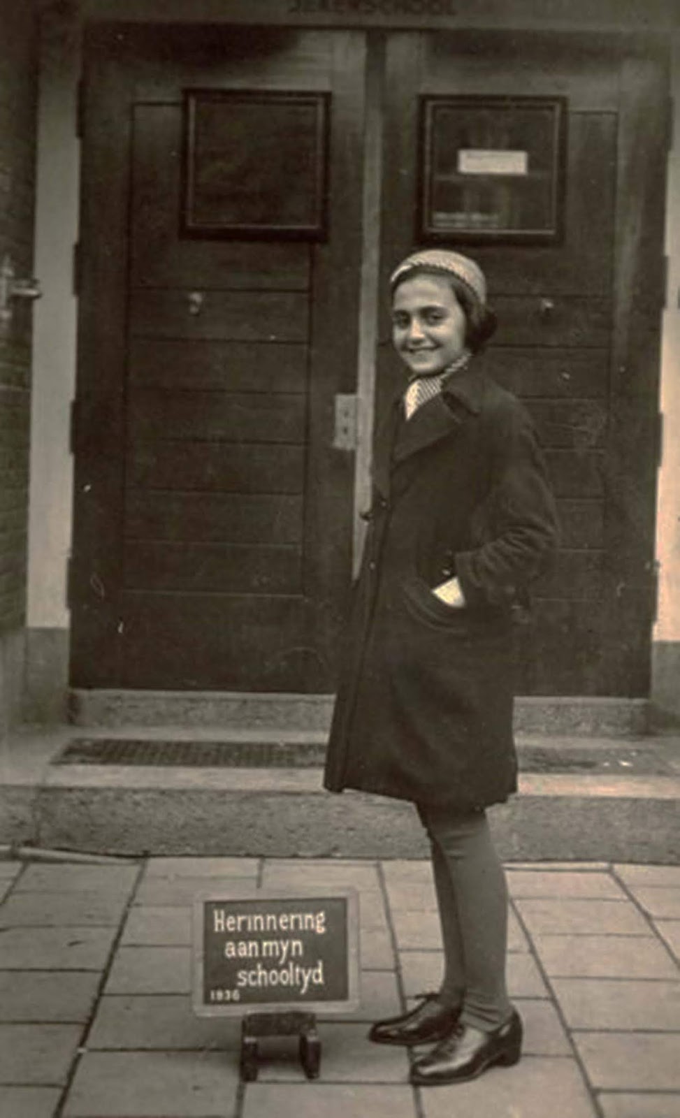 Margot Frank, the older sister of Anne Frank, standing in front of the doorway of the Jeker school in Amsterdam with a plaque at her feet that says 'memory of my school year 1936' in Dutch.