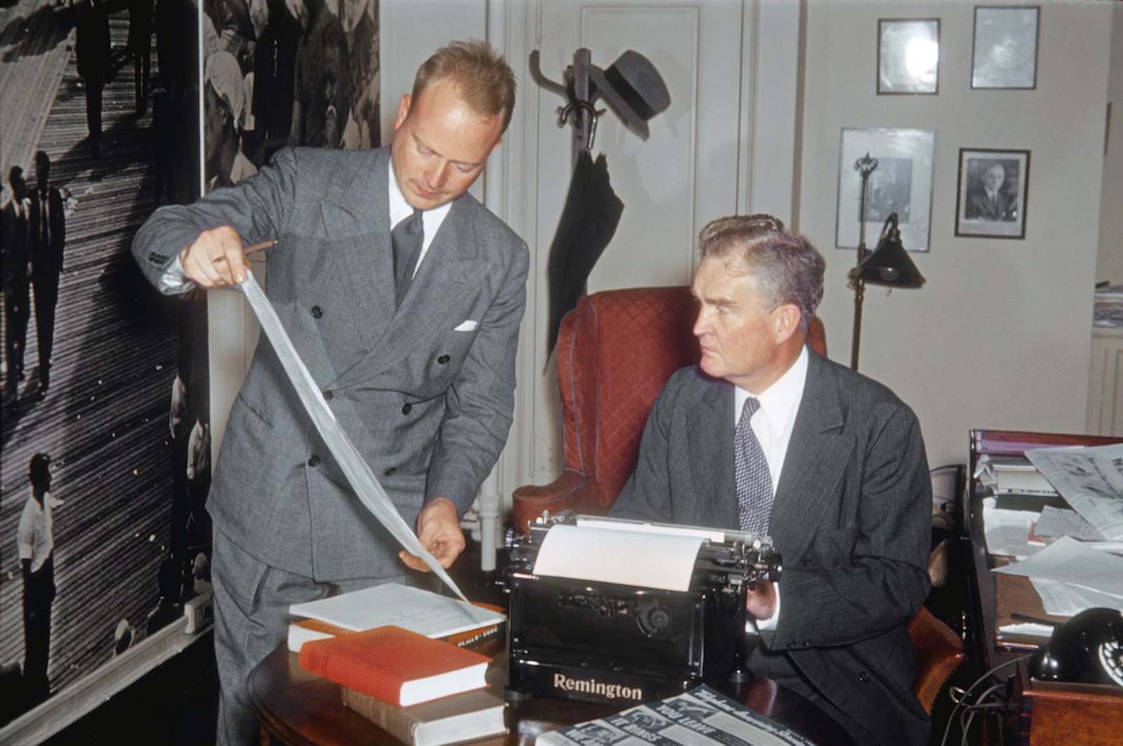 Two men review an ad spread for a telephone answering machine, one sits behind a classic Remington typewriter.