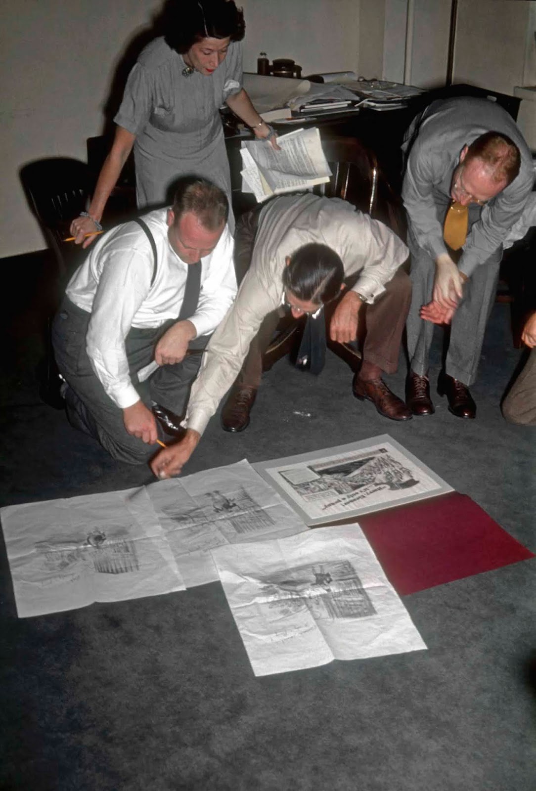 A female secretary clutches a multitude of notes, as three men examine a range of illustrations.