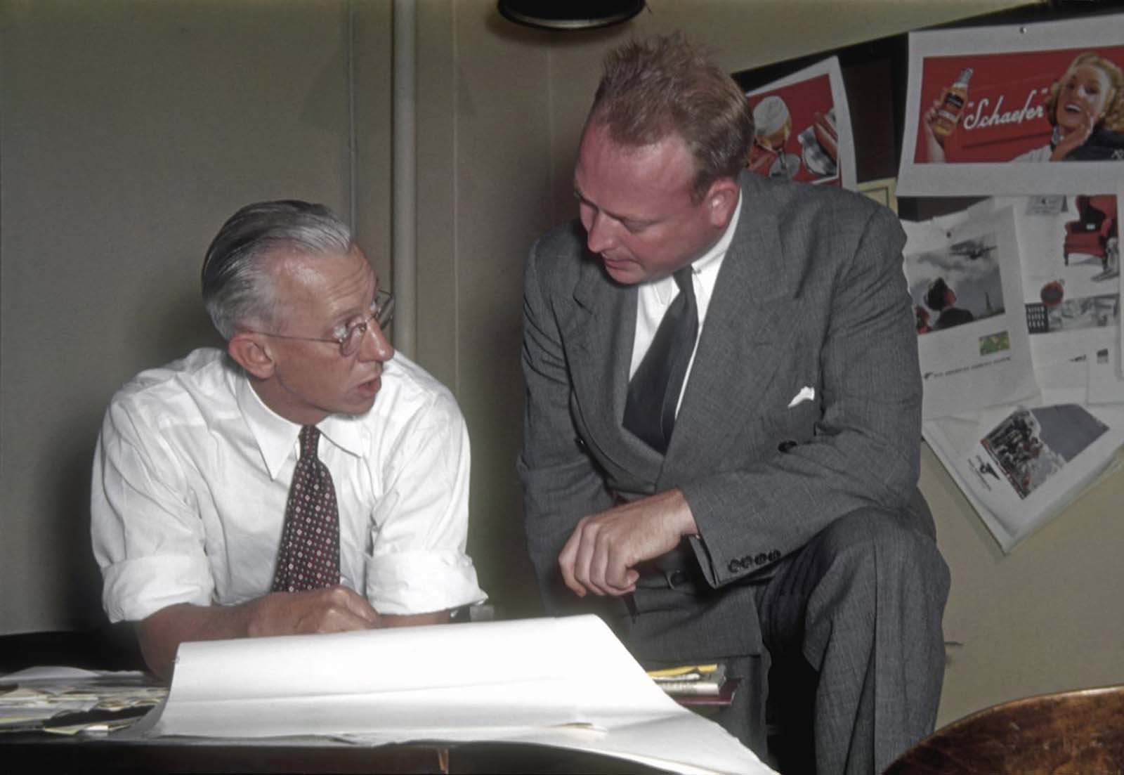 Two ad men discuss a campaign, a pinboard behind them is covered with a range of spreads, including one for Schaefer beer.
