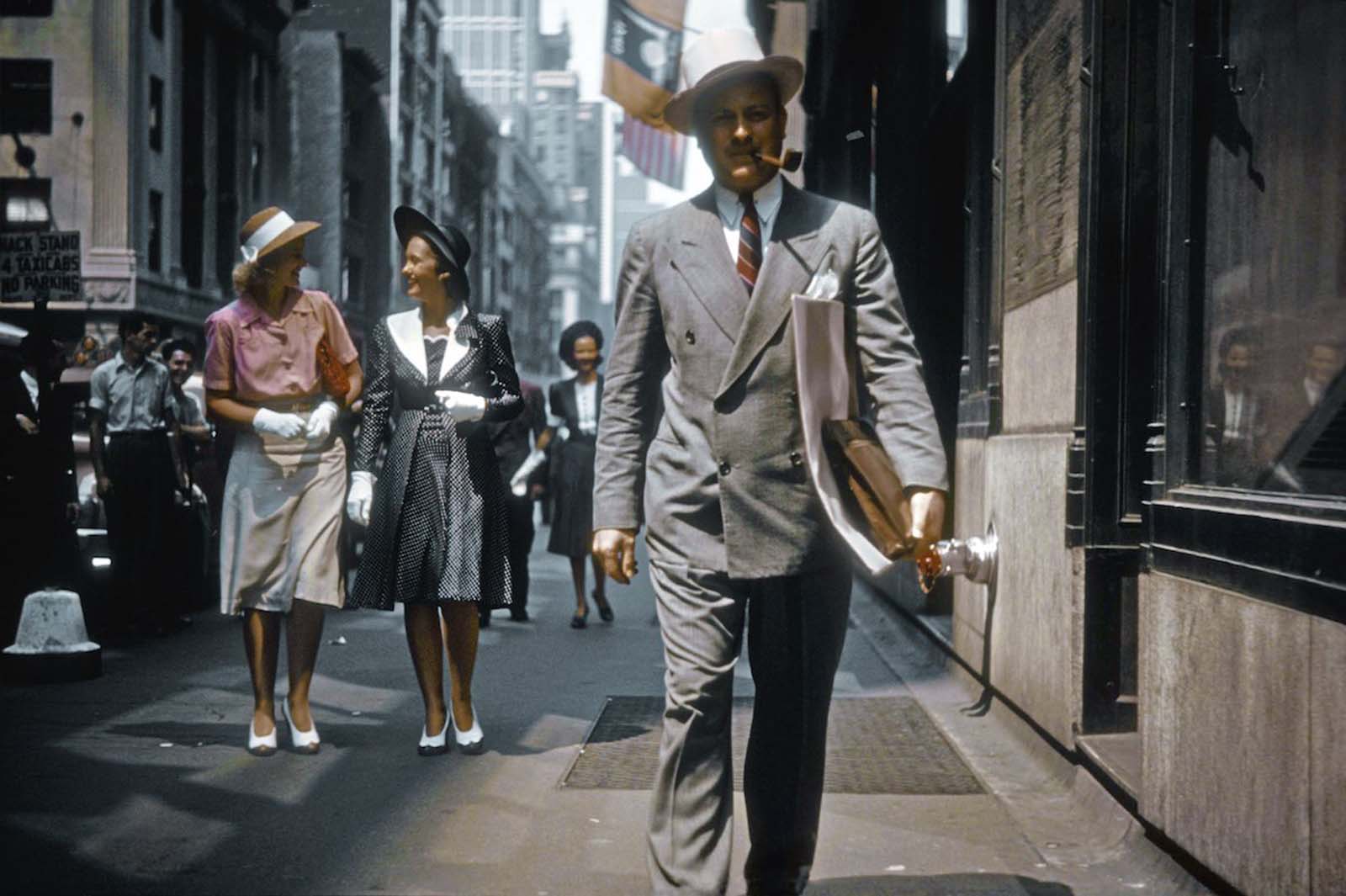 A man walks down Madison Avenue on a clear, warm day. Behind him, three women are wearing white gloves.