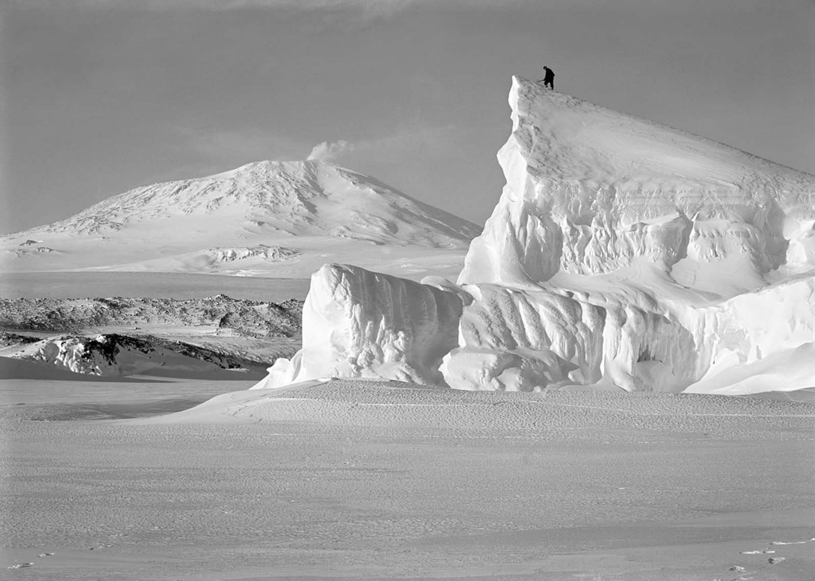 A man stands atop the Matterhorn Berg with active volcano Mt. Erebus in the background. October 8, 1911.
