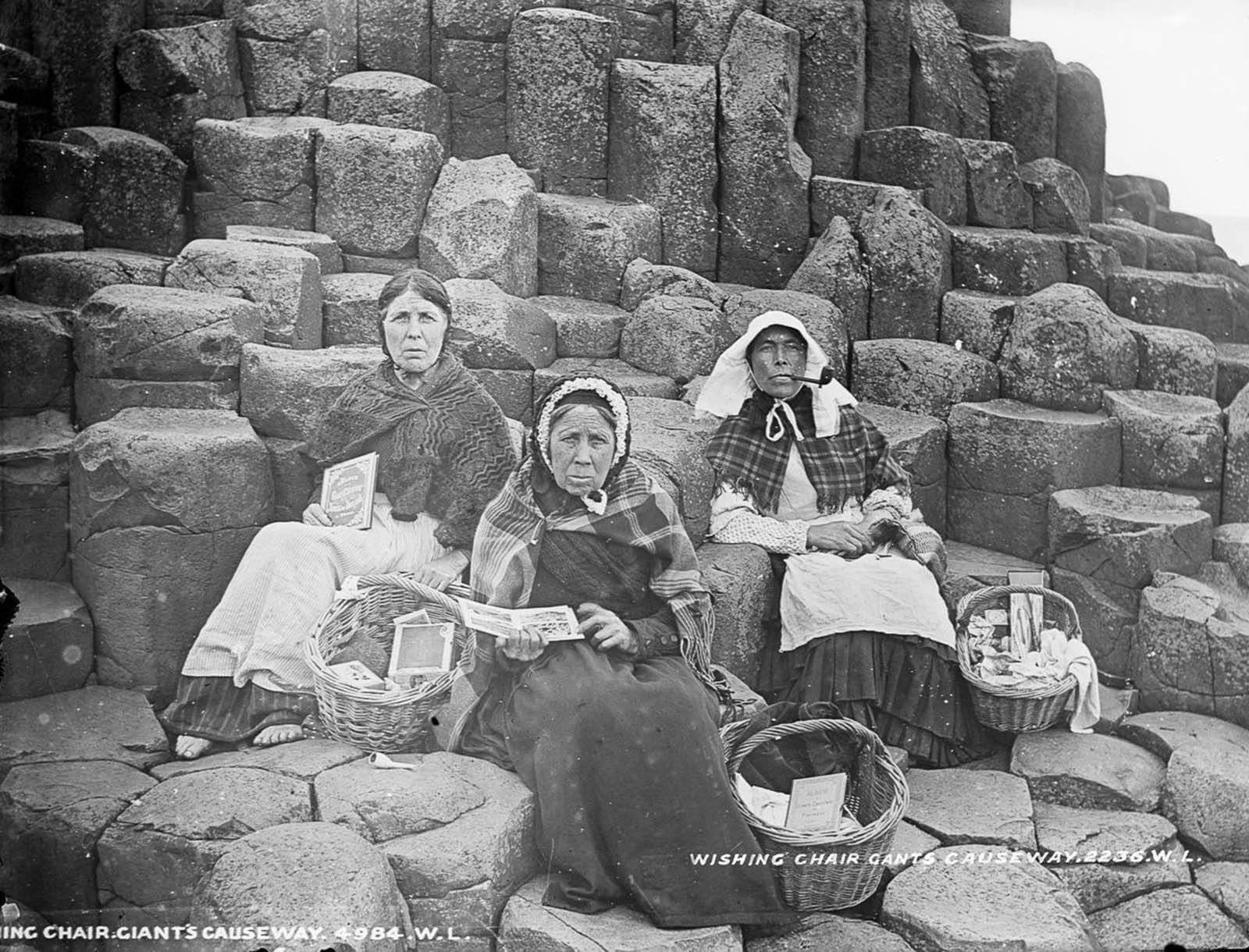 Women sell tourist trinkets and books on Fionn Mac Cumhaill's Wishing Chair at the Giant's Causeway, County Antrim. 1900.