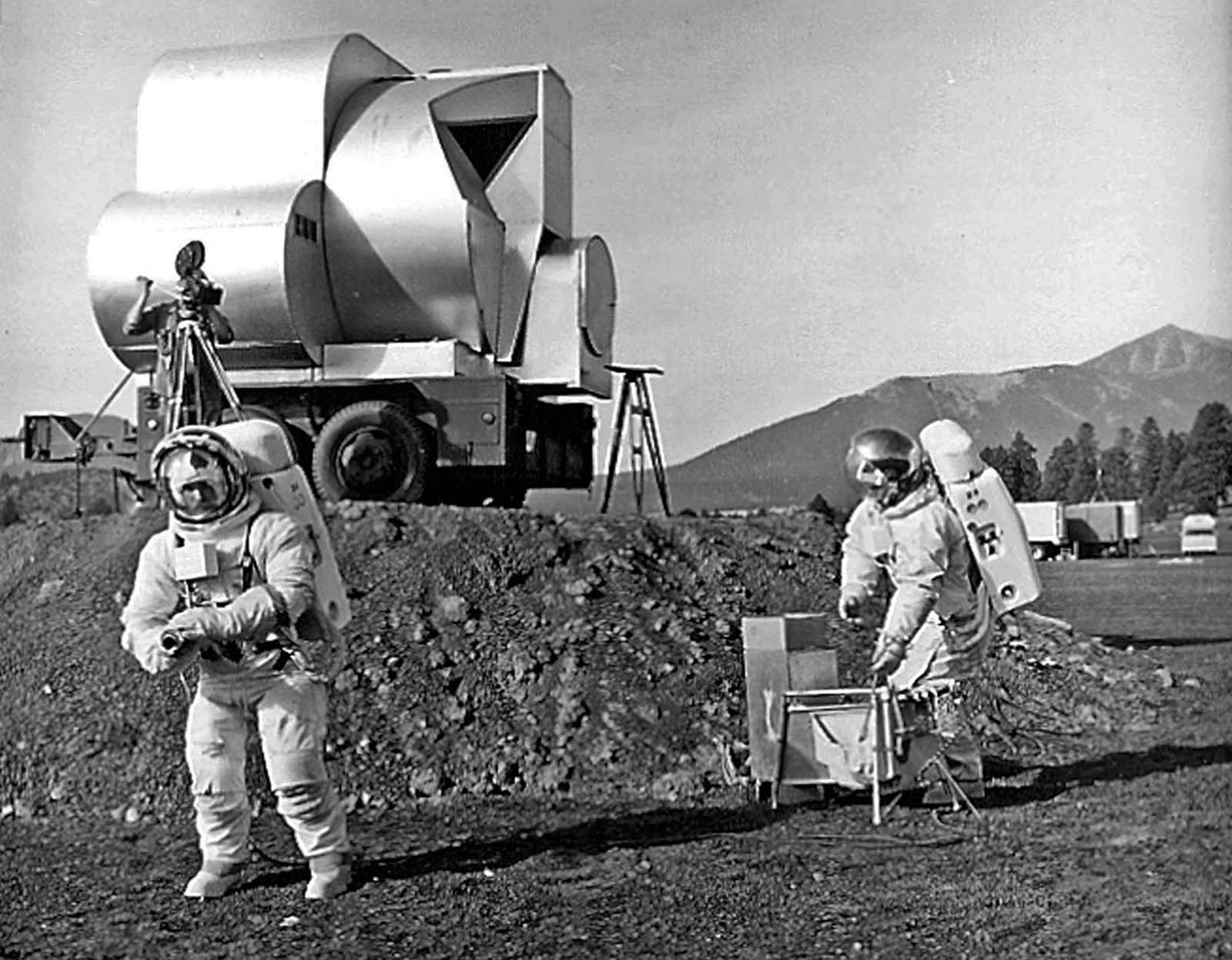 The USGS geologists Tim Hait and David Schleicher work in spacesuits during an early Apollo field test at Cinder Lake Crater Field #1, with a simulated lunar-module ascent stage mock-up parked on an earthen ramp in the background.